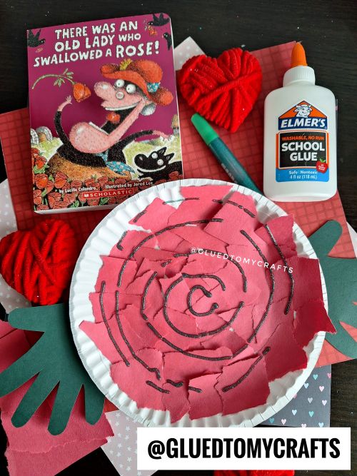 A children’s craft project is displayed with a paper plate rose made from torn pink paper, a bottle of Elmer’s glue, a green marker, red yarn hearts, and the book There Was an Old Lady Who Swallowed a Rose!—a perfect Valentine’s Day craft idea for kids.