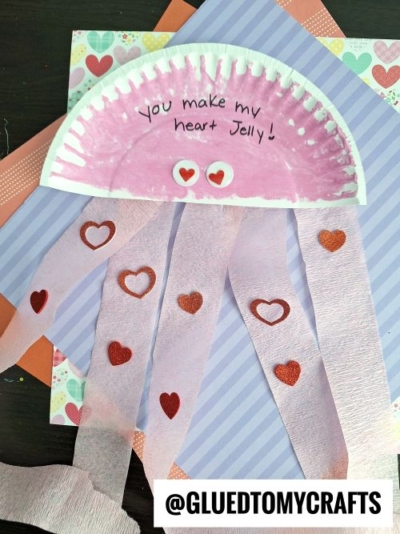 A pink paper plate jellyfish craft with "You make my heart jelly!" written on it—perfect for valentine's day craft ideas for kids. The jellyfish features crepe paper tentacles decorated with heart stickers and a colorful patterned background.