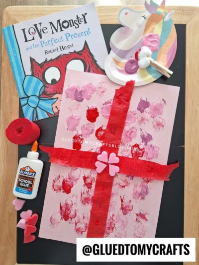 A pink Valentine's Day craft with red paint, heart stickers, and ribbon sits on a tray, alongside an Elmers glue bottle, the book Love Monster and the Perfect Present—a fun setup for valentine's day craft ideas for kids—with a unicorn paint palette and brushes.