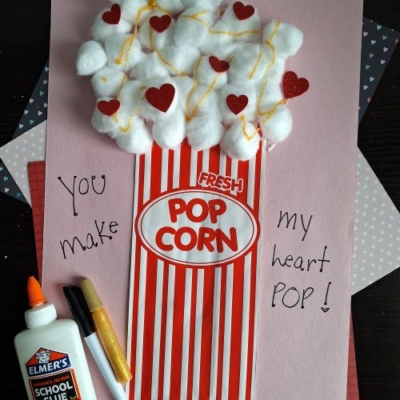 A handmade Valentine’s Day card resembling a popcorn box with cotton ball popcorn, red hearts, and yellow lines—perfect for valentine's day craft ideas for kids. “You make my heart pop!” is written on it. School glue and pens are beside the card.