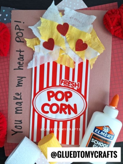 A handmade Valentine’s card features a popcorn box with red stripes, yellow and white tissue “popcorn” with red hearts, a love note reading “You make my heart POP!”, and a bottle of Elmer’s glue—perfect for Valentine’s Day craft ideas for kids.