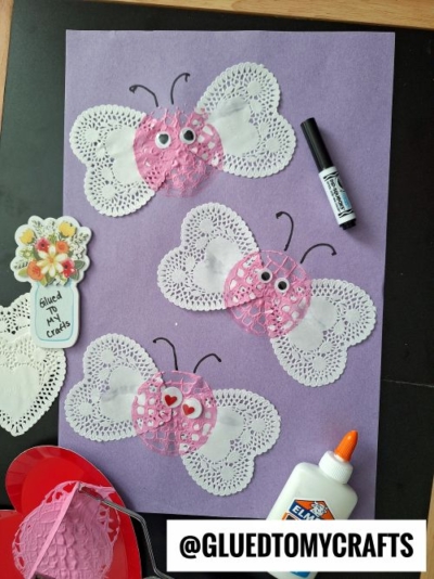 Three butterfly crafts made from white heart-shaped paper doilies for wings and pink doilies for bodies, arranged on a purple background. The butterflies have drawn antennae and googly eyes or heart-shaped stickers. Surrounding the craft are a glue bottle, marker, extra doilies, and a sticker reading "Glued To My Crafts." The image is branded with "@GLUEDTOMYCRAFTS."