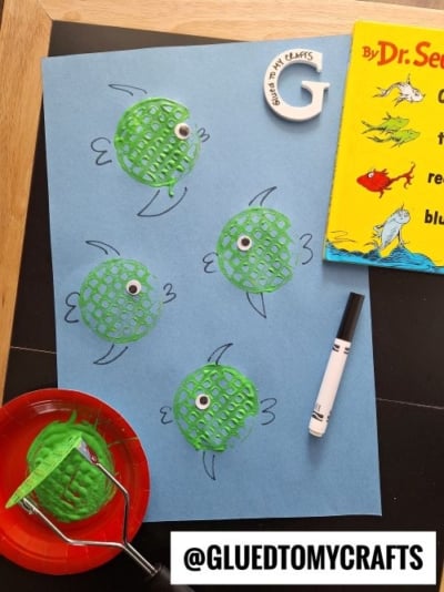 A fish-themed craft inspired by Dr. Seuss, featuring green potato masher-stamped fish with googly eyes and black marker-drawn fins on a blue paper. A red plate with green paint and a potato masher are visible, along with a Dr. Seuss book and a white "G" cutout with "GLUED TO MY CRAFTS" written on it.