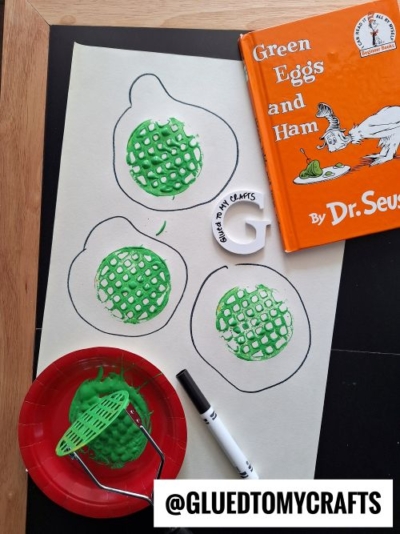 A craft inspired by "Green Eggs and Ham" by Dr. Seuss, featuring a white paper with three black marker-drawn egg outlines filled with green textured paint, mimicking green eggs. A red plate with green paint and a potato masher is visible, along with the book and a white "G" cutout with "Glued TO MY CRAFTS" written on it.