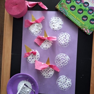 A purple sheet with potato masher stamped unicorn faces, decorated with pink crepe paper manes and gold paper horns, sits next to a book, a roll of crepe paper, and a purple plate with paint. @GLUEDTOMYCRAFTS is labeled.