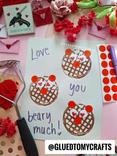 A handmade card says Love you beary much! with three bear faces made from brown waffle prints and red stickers—one of the cutest Valentine's Day craft ideas for kids—surrounded by craft tools, fake flowers, and red sticker sheets on a pink surface.