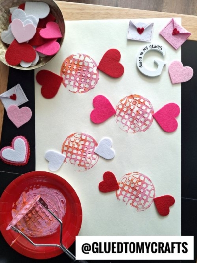 A Valentine's Day craft showing candy designs made from textured pink and orange painted circles with felt hearts as wrappers. The candies are displayed on a light surface, surrounded by crafting materials like a red plate with paint, a textured roller, a metallic bowl of felt hearts, and small felt envelopes. The branding "@GLUEDTOMYCRAFTS" is visible in the corner.