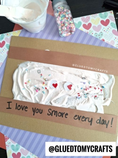 A handmade card with a brown strip reading I love you Smore every day! features white puffy paint topped with colorful sprinkles and hearts, making it one of the sweetest Valentine's Day craft ideas for kids. Patterned paper and crafting supplies sit nearby.