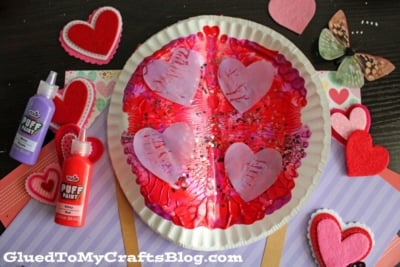A paper plate painted with red and pink puff paint, decorated with tissue paper hearts and glitter. Surrounded by felt heart cutouts, a butterfly embellishment, and bottles of red and purple puff paint.