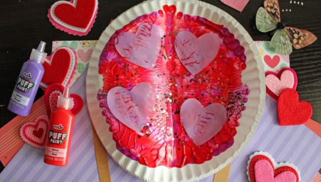A paper plate painted with red and pink puff paint, decorated with tissue paper hearts and glitter. Surrounded by felt heart cutouts, a butterfly embellishment, and bottles of red and purple puff paint.