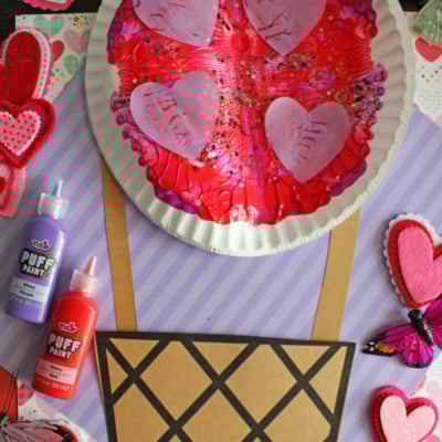 A colorful craft featuring a paper plate decorated with pink heart shapes and red paint as a hot air balloon, with a brown paper basket, surrounded by felt hearts and butterfly decorations. Text reads: Paper Plate & Paint Splat Hot Air Balloon Craft.