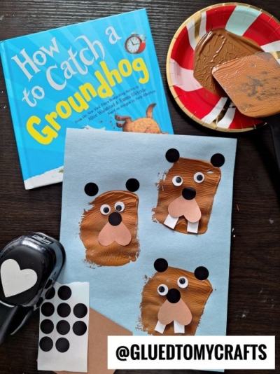 A childrens book titled How to Catch a Groundhog, a paper with four painted groundhog faces, a plate with brown paint, a brush, a paper punch, and black dot stickers are arranged on a table.