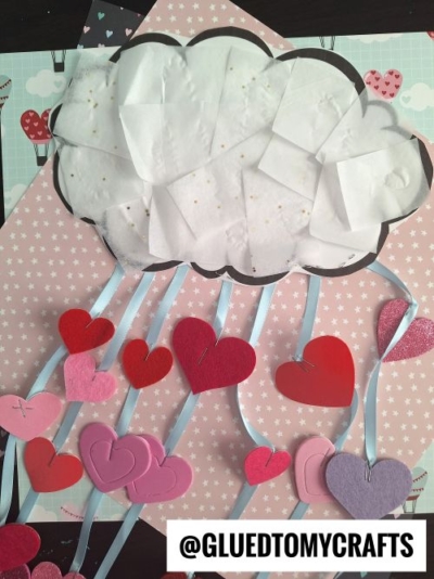 A cloud made of white paper is attached to a pink polka-dot background. Ribbons hang down from the cloud with red, pink, and purple hearts attached, creating a rain effect. @GLUEDTOMYCRAFTS is written at the bottom.