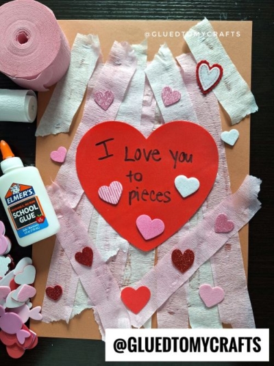 A perfect Valentine’s Day craft idea for kids: brown paper adorned with white and pink crepe paper strips, a red heart saying “I love you to pieces,” surrounded by foam hearts, pink paper hearts, a glue bottle, and a roll of crepe paper.