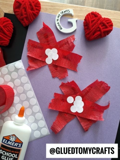 A fun Valentine's Day craft idea for kids: two red crepe paper bows with white adhesive dots on a purple background. Red yarn hearts, a bottle of Elmer’s glue, and a sticker sheet complete the scene. @GLUEDTOMYCRAFTS at the bottom.