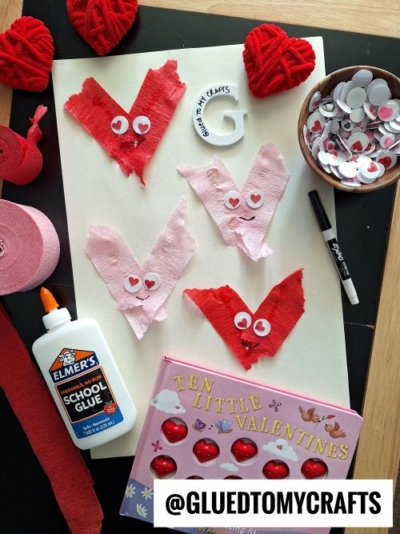A flat lay of a Valentine’s Day craft project features red and pink tissue paper hearts with googly eyes, a glue bottle, foam hearts, a marker, children’s book, and @GLUEDTOMYCRAFTS—perfect for fun Valentine's Day craft ideas for kids.