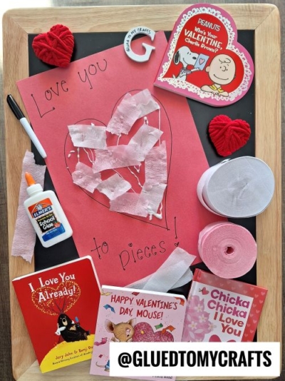 A Valentines Day craft display on a chalkboard features a red paper heart collage, glue, yarn hearts, two rolls of crepe paper, and several Valentine-themed children’s books—perfect valentine's day craft ideas for kids. @GLUEDTOMYCRAFTS is written at the bottom.
