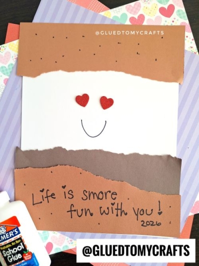A handmade card with torn brown and tan paper layers resembling a smore makes a cute addition to valentine's day craft ideas for kids. It features a smiling face with heart eyes, the phrase "Life is smore fun with you! 2020," plus glue and papers nearby.
