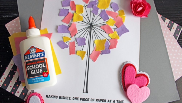 Flat lay of a torn paper dandelion craft featuring a white sheet with a black dandelion outline. Torn yellow, pink, and purple paper pieces are glued to the dandelion head, simulating seeds. Surrounding the craft are Elmer's glue, felt hearts, paper flowers, and patterned scrapbook paper. The text 'MAKING WISHES, ONE PIECE OF PAPER AT A TIME.' and 'GluedToMyCraftsBlog.com' are printed on the white sheet.