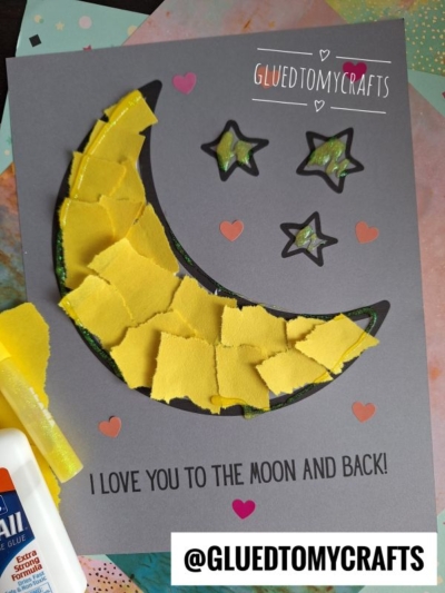 A handmade card features a torn paper moon crafted from yellow paper, surrounded by four black-outlined stars. Text reads, I love you to the moon and back! Glue and paper scraps are visible nearby.