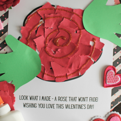 A handmade rose Valentine’s Day craft made from red paper pieces with glitter on a printed sheet that reads, A rose that wont fade! Green paper leaves, Elmer’s glue bottle, and heart decorations surround the craft.