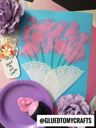 A handmade craft featuring pink painted flowers with green stems on blue paper, doily pieces as vases, and purple artificial flowers. Craft supplies and a label reading @GLUEDTOMYCRAFTS appear in the image.