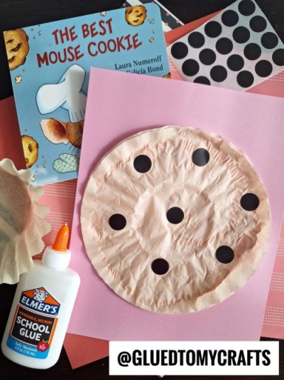 A children's craft project inspired by the book "The Best Mouse Cookie" by Laura Numeroff. The craft features a crumpled light brown coffee filter decorated with black circular stickers to resemble a chocolate chip cookie. The setup includes the book, a bottle of Elmer's glue, and colorful background papers in pink and reddish tones.