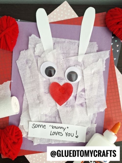 A handmade bunny craft for Valentine's Day, featuring a textured face made from white crepe paper, googly eyes, and a red heart-shaped nose. The bunny has white ears and is set against a colorful background of patterned papers in purple, red, and pink. Craft supplies like a glue bottle and crepe paper roll are visible nearby.