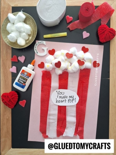 A Valentines Day craft on pink paper shows a popcorn bucket made with red crepe paper strips and cotton balls, with a heart note reading You make my heart POP! Surrounded by glue, yarn hearts, cotton balls, and craft supplies.
