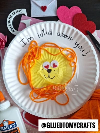 A paper plate craft features a yellow lion face made from paper and orange yarn for the mane, with heart stickers for eyes. Im wild about you! is written above. Craft supplies and @GLUETOMYCRAFTS label are visible.