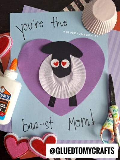 A handmade card featuring a sheep made from a white cupcake liner, black paper, and googly eyes with heart-shaped pupils. The sheep is placed on a purple heart background with the message "You're the baa-st Mom!" The card is surrounded by craft supplies like glue, scissors, and felt hearts, set against a colorful backdrop of patterned papers.