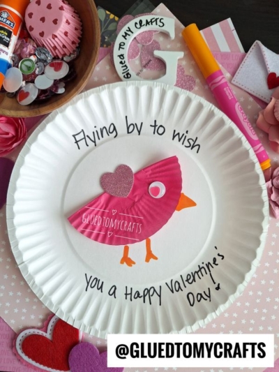 A paper plate craft features a pink bird made from cupcake liners and paper, with Flying by to wish you a Happy Valentine’s Day handwritten around it. Craft supplies and decorations are scattered in the background.