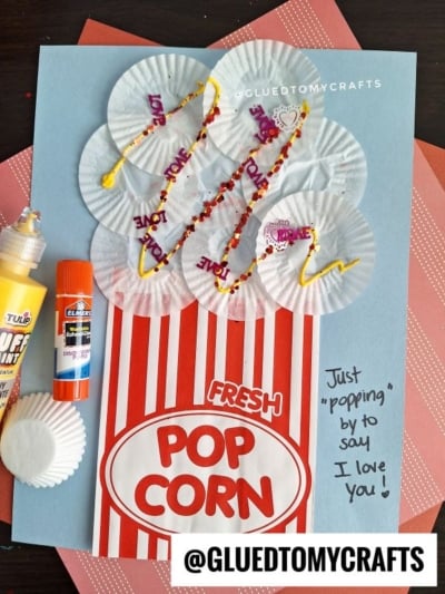 A handmade popcorn craft with cupcake liners as popcorn, red striped paper as the popcorn box, POP CORN label, yellow puffy paint, and Just popping by to say I love you! handwritten on blue paper.