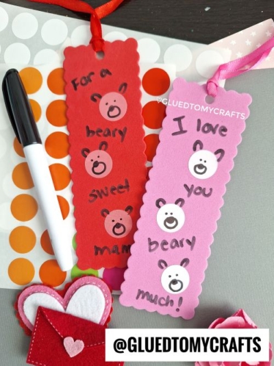 Two handmade bookmarks, one red and one pink, with scalloped edges, bear face stickers, and sweet messages like "I love you beary much!" The bookmarks are surrounded by a marker, polka-dotted papers, and felt heart embellishments. Branding from "Glued To My Crafts" is included.