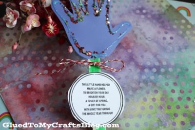 A colorful handprint craft made from purple paper with glitter edges sits on a multicolored polka dot background. A round tag with a poem is attached below, tied with red and white string. Flowers are visible in the corner.