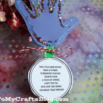 A colorful handprint craft made from purple paper with glitter edges sits on a multicolored polka dot background. A round tag with a poem is attached below, tied with red and white string. Flowers are visible in the corner.