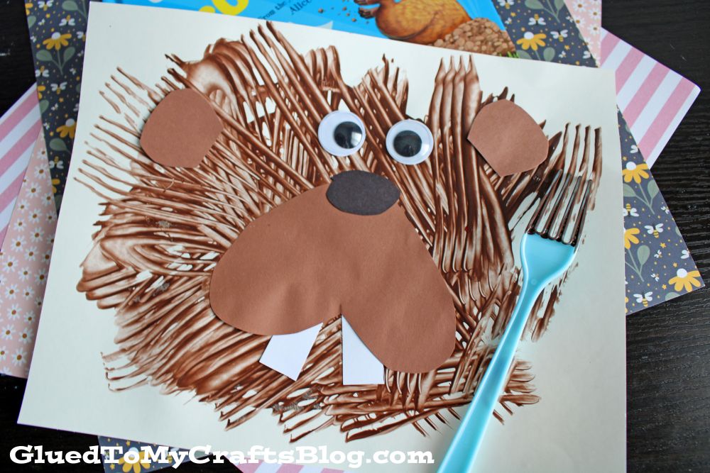 A children’s craft of a beaver made with brown paint strokes, googly eyes, and paper shapes for the nose, teeth, and ears. A blue plastic fork used for painting is beside it.