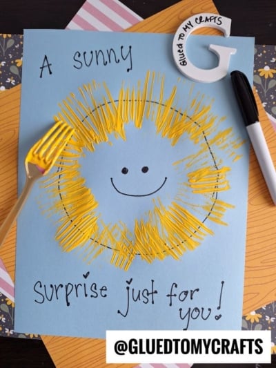 A blue paper with a hand-drawn sun and face, yellow paint sun rays made with a fork, the phrase A sunny surprise just for you!, a white pen, a decorated G, and patterned paper beneath. @GLUETOMYCRAFTS tag at the bottom.
