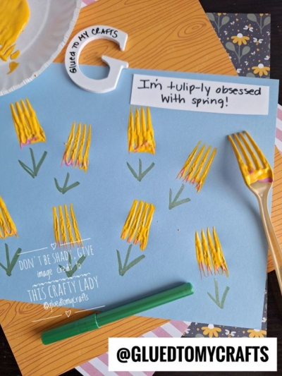 A blue sheet of paper displays yellow tulip flowers painted with a fork, with green stems drawn in marker. A note reads, I’m tulip-ly obsessed with spring! Craft supplies are scattered around.