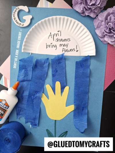 A craft project features a yellow paper flower on blue paper, with a white paper plate half-circle above it labeled April showers bring may flowers! Blue crepe paper strips hang down as rain. A glue bottle and supplies are nearby.