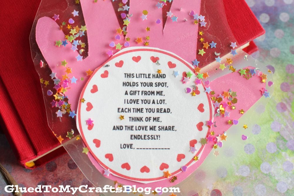 A pink hand-shaped bookmark with red heart stickers and star confetti surrounds a round note that reads a heartfelt message about love and reading. The image is from GluedToMyCraftsBlog.com.