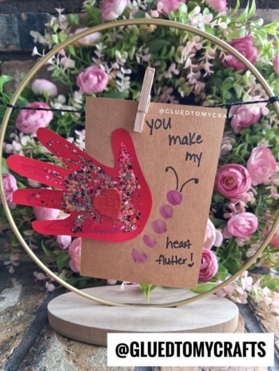 A handmade card with a red handprint butterfly and glitter, clipped to a stand, reads You make my heart flutter! Pink flowers are in the background. @GLUEDTOMYCRAFTS is written at the bottom.