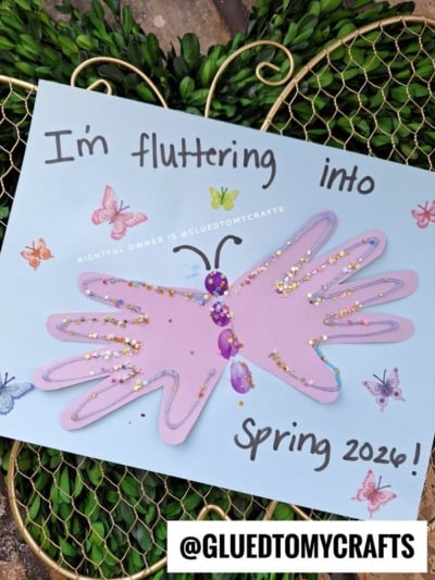 A handmade butterfly craft made from two pink handprints decorated with glitter and gems is glued to blue paper with colorful butterfly illustrations and the words “I’m fluttering into Spring 2026!”.
