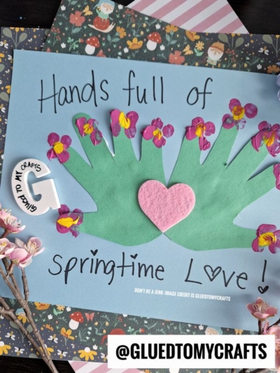 A craft project features two green handprints with pink and yellow flower fingertip accents, a pink felt heart in the center, and the words “Hands full of springtime love!” written on blue paper.