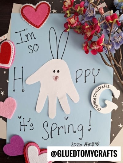 A spring craft featuring a blue paper with a white handprint bunny, handwritten Im so Hoppy its Spring!, felt hearts, a felt C-shape labeled GluedToMyCrafts, and artificial flowers.