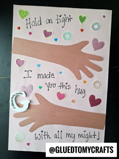 A pink card with two cut-out paper arms, heart and flower stickers, and handwritten text: Hold on tight, I made you this hug with all my might! A white “G” with “@GLUEDTOMYCRAFTS” is in the lower left corner.