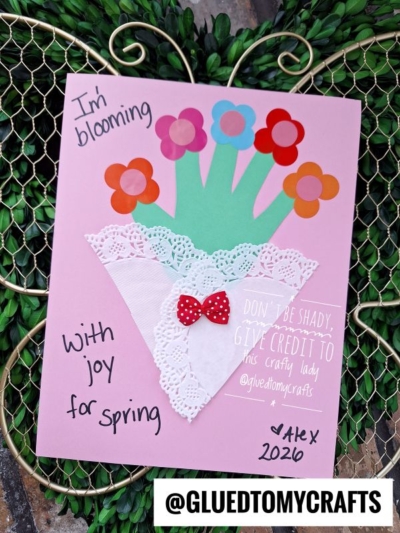 A handmade card with colorful paper flowers in a white paper doily vase, decorated with a red bow. Text reads Im blooming with joy for spring. The card is signed “Alex 2020” and tagged @gluedtomycrafts.