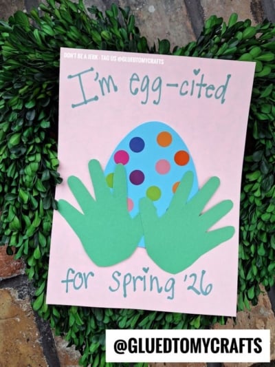 A pink card with a colorful polka-dot egg and two green handprint cutouts reads, Im egg-cited for Spring 26. The card is placed on greenery, with @GLUEDTOMYCRAFTS at the bottom.