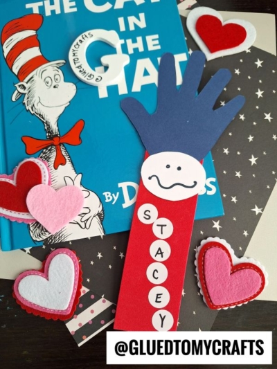A handmade bookmark shaped like a blue hand with a red arm and white face labeled STACEY lies on a Cat in the Hat book, surrounded by felt hearts on star-patterned paper. @GLUEDTOMYCRAFTS is written at the bottom.