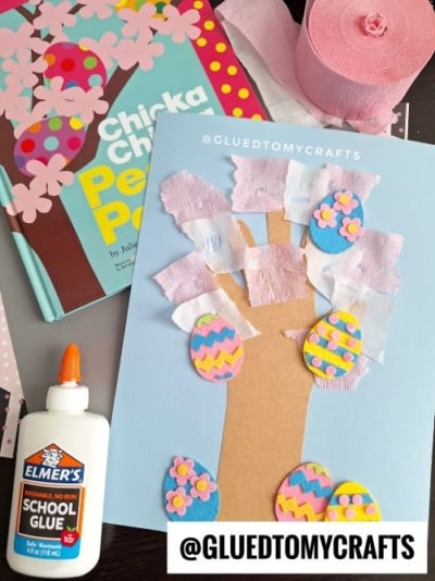 A spring or Easter-themed craft showing a cherry blossom tree made from a brown paper trunk and branches, decorated with torn pink and white crepe paper as blossoms and felt Easter eggs. The craft is displayed on a light blue background with a roll of pink crepe paper, a glue bottle, and a children's book titled "Chicka Chicka Peep Peep" nearby. The watermark "@GLUEDTOMYCRAFTS" is visible.